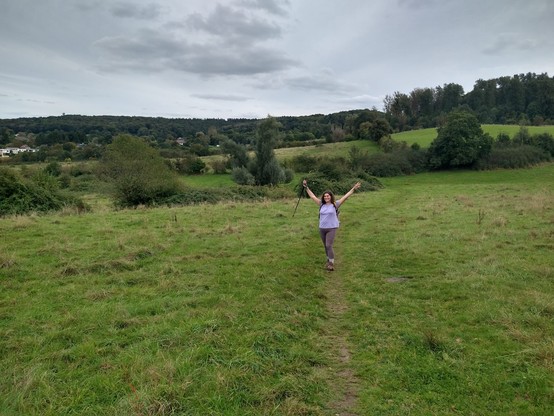 Me, standing in a trail passing a pasture on a hill. I have my arms raised and have a walking stick in my right hand. Around the pasture are bushes, and the area is hilly with forests in the distance. I am wearing a light purple shirt and brownish hiking leggings and hiking boots.