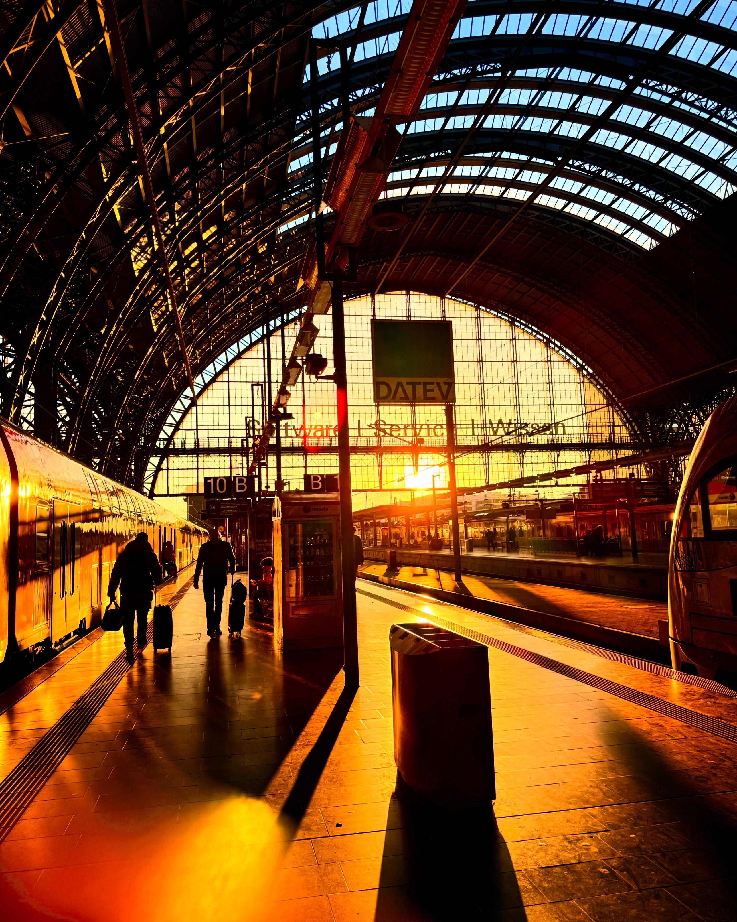 Sonnenuntergang am Bahnhof – Silhouetten von Reisenden im goldenen Licht.


Die tief stehende Sonne strahlt durch das große Glasdach eines Bahnhofs und taucht die Szene in warmes, goldenes Licht. Lange Schatten fallen auf den Bahnsteig. Zwei Personen mit Rollkoffern gehen entlang eines haltenden Zuges, während im Hintergrund weitere Reisende und Bahnsteigschilder zu sehen sind. Die beeindruckende Architektur des Bahnhofs mit geschwungenen Stahlträgern und Glasfenstern wird von der untergehenden Sonne dramatisch beleuchtet.